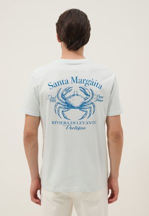 Persona che indossa una maglietta bianca con un'illustrazione di un granchio blu e testo in italiano su Santa Margàita e Portofino sulla schiena.