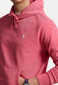 Rosa Kapuzenpullover aus Baumwolle mit Kängurutasche, Kordelzugkapuze und einem weißen, gestickten Logo auf der Brust. Weiche Textur und lässiges Design.