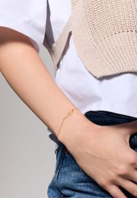 Złota bransoletka na nadgarstku, z delikatnym wzorem łańcuszka. Tekstura bransoletki jest gładka, a jej estetyka minimalistyczna.