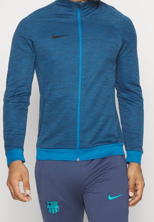 Mann trägt eine blaue Nike-Reißverschlussjacke und dunkelblaue Hose mit FC Barcelona- und Nike-Logos.