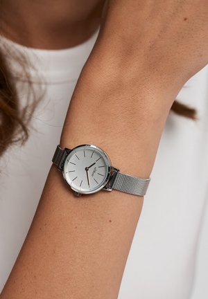 Sølv håndledsur med rund urskive, mesh-rem og minimalistisk design. Skiven har tynde time-markører og en glat, reflekterende overflade.