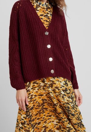 Vínový pletený cardigan s výstřihem do V, se čtyřmi stříbrnými knoflíky a texturovanými vzory. Nosený přes žluto-hněd dresses s vzorem.