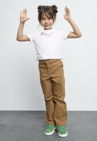 Vit t-shirt med sidofästen och en ljusgrå grafisk bild, kombinerat med beige byxor och gröna sneakers. Barnet har händerna uppe och ler.