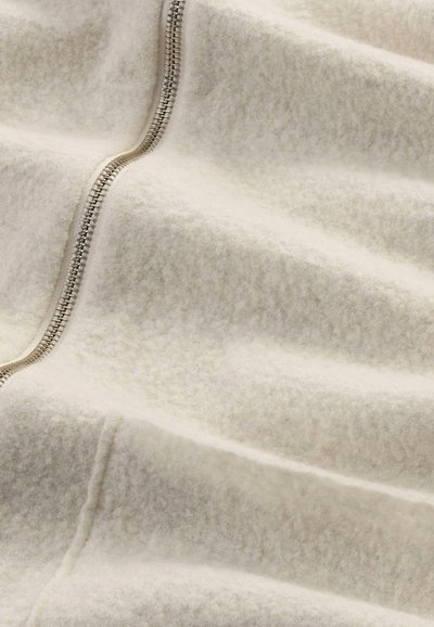 Cremfarbende Fleece-Decke mit glatter Textur und einem sichtbaren silbernen Reißverschluss entlang einer Kante, die sanfte, sanft geschwungene Falten zeigt.