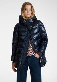 Giacca a vento blu navy con finitura lucida, cappuccio e design patchwork sotto. Presenta una chiusura con zip e tasche laterali.