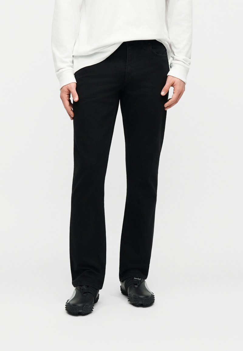 Homme portant un pantalon droit noir, des chaussures noires et une chemise blanche à manches longues, debout devant un fond blanc.