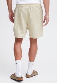 Light beige shorts with an elastic waistband and a back pocket. The fabric has a subtle texture. Worn with white socks and sandals.