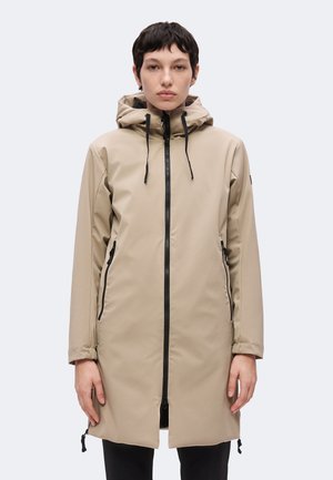 Ung person med kort mørkt hår iført en beige hættetrøje med lynlås og sorte lynlås samt sidelommer, stående mod en ensfarvet baggrund.