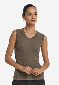 Kobieta z ciemnymi włosami upiętymi w kok, ubrana w bezrękawnik w kolorze taupe z materiału prążkowanego oraz złote kolczyki w kształcie obręczy, delikatnie złożone ręce przed sobą.
