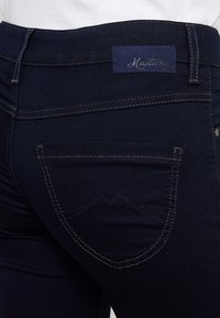 Granatowe dżinsy o wysokim stanie, z kontrastowym przeszyciem i metką z logo "Mustang" na tylnej części pasa.