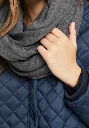 Femme portant une veste matelassée bleu marine et une grosse écharpe en tricot gris, tenant l'écharpe près du cou avec la main droite.