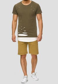 Homme portant un t-shirt vert olive usé par-dessus un sous-shirt blanc, un short beige et des baskets blanches, debout devant un fond gris.