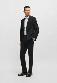 Completo nero con giacca aderente e pantaloni a gamba dritta, abbinato a una camicia bianca. Tessuto liscio con una leggera trama. Scarpe nere.