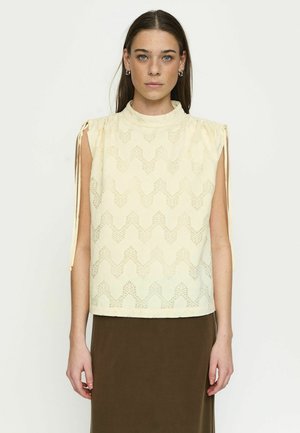 Kobieta o prostych brązowych włosach, ubrana w kremową bluzkę bez rękawów z marszczonymi ramionami i brązową spódnicę, zwrócona przodem.