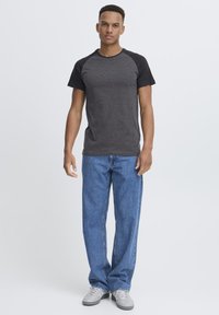 Camiseta raglán de manga corta en gris oscuro con mangas negras, combinada con jeans rectos de pierna ligera en azul y zapatos grises. Fondo liso.