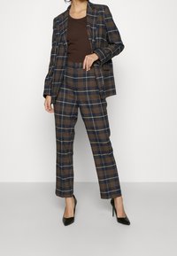 Merretti ja ruskeat ruudulliset puvut, joissa on muotoiltu bleiseri ja slim fit -housut, valmistettu tekstuuripinnoitteisesta kankaasta, yhdistetty mustiin korkokenkiin.