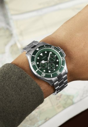 Srebrna kovinska ročna ura z zelenim številčnico in kronografskimi funkcijami, nošena na zapestju preko zelene majice, v ozadju zamegljena karta.