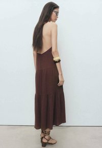 Robe maxi sans manches, marron, avec un dos décolleté, un design en étages et un tissu texturé. Associée à des sandales brunes à lanières et des bracelets.