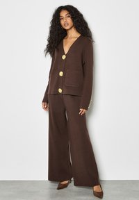 Cardigan lavorato a maglia marrone con scollo a V, grandi bottoni dorati, due tasche frontali, abbinato a pantaloni larghi coordinate e tacchi a punta marroni.