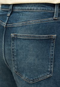 Κοντινή λήψη της πίσω τσέπης και των λεπτομερειών ραφής σε μπλε τζιν τζιν φόρεμα, φορε μένα με μπεζ πλεκτό τοπ.