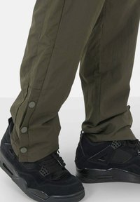 Olijfgroene broek met drukknoopjes aan de onderkant, gecombineerd met zwarte sneakers met een gestructureerd mesh en suède bovenmateriaal.