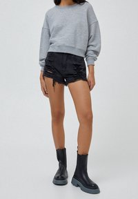 Mujer con sudadera gris, pantalones cortos de mezclilla negra desgastados y botas negras gruesas con suelas grises gruesas, de pie sobre un fondo liso.