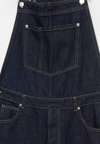 Temno modri džins overali z srebrnim okovjem, oranžnim šivanjem, sprednjim žepom in stranskima žepoma ter teksturirano površino.