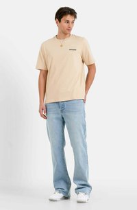 Camiseta de algodón beige con cuello redondo y un logo verde, combinada con jeans de corte relajado azul claro y zapatillas blancas.