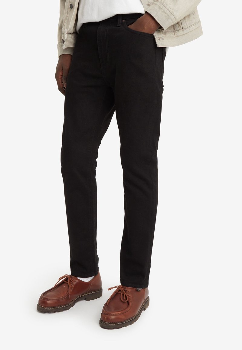 Homme portant un pantalon noir slim, des chaussures en cuir marron à lacets, des chaussettes blanches, une veste légère, main dans la poche, debout sur fond blanc.