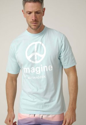 Hombre que lleva una camiseta azul claro con un símbolo de la paz blanco y la palabra "imagine" encima de unos pantalones cortos multicolor, mirando hacia abajo.