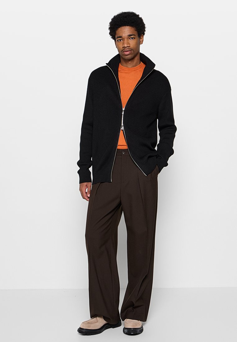 Schwarzer Reißverschluss-Pullover mit gerippter Textur, getragen über einem leuchtend orangen Hemd. Kombiniert mit lockeren braunen Hosen und hellbraunen Schuhen.