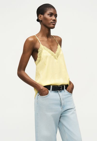 Γυναίκα με κοντά μαλλιά που φοράει κίτρινο τοπ με δαντέλα, μπουστάκι, μπουστωμένο σε ανοιχτό μπλε ψηλόμεσα τζιν με μαύρη ζώνη.