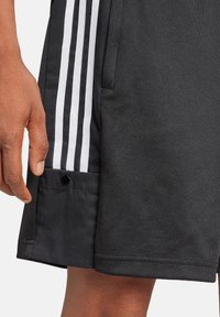 Schwarze Sportshorts mit drei weißen Streifen an der Seite, einer Seitentasche und einem kleinen schwarzen Druckknopf-Detail am Saum.
