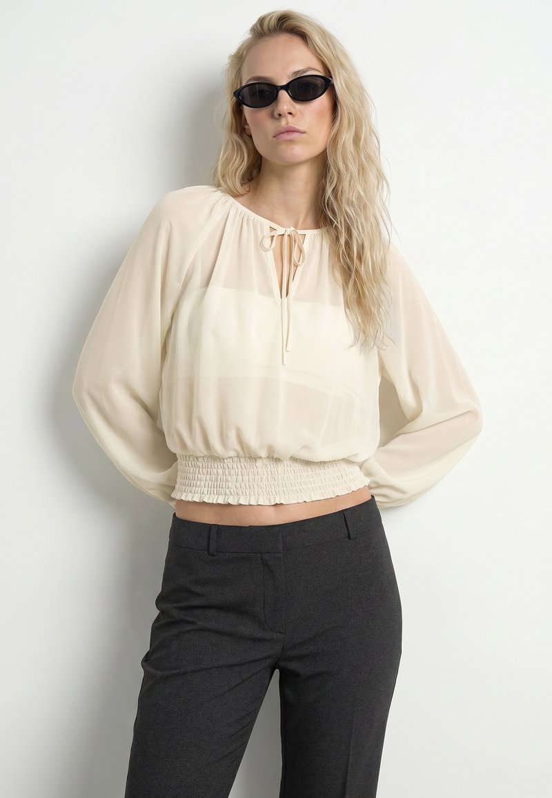 Femme blonde portant des lunettes de soleil noires œil de chat, un chemisier crème transparent avec une taille smockée, et un pantalon gris foncé sur un fond clair uni.