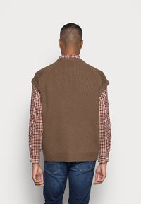 Gilet en tricot marron avec épaules raglan, tissu texturé et design côtelé, porté sur une chemise à manches longues à carreaux, associé à un jean bleu.