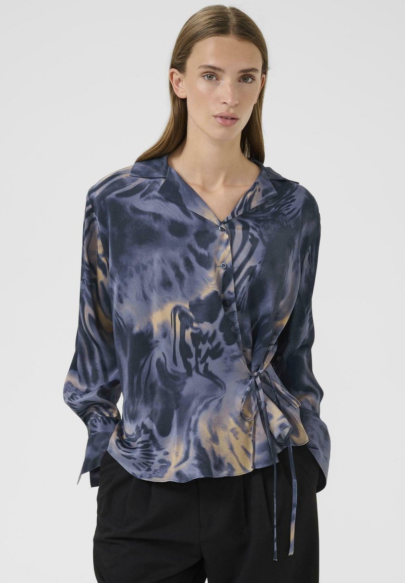 Wikkelblouse van soepel materiaal met een blauw-gouden abstract patroon. Afgewerkt met een kraag en een strikdetail op de taille. Lange mouwen.