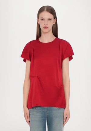 Haut rouge à manches courtes en tissu lisse, avec un col rond et de légères plis près des poches. Coupe légèrement ample, associé à un jean bleu clair.