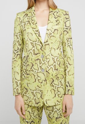 Vrouw draagt een geelgroene blazer met slangenprint en twee knopen over een witte top, gecombineerd met een bijpassende broek met slangenprint.