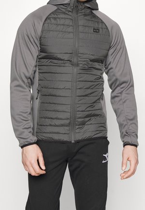 Homme portant une veste matelassée grise à capuche avec des manches gris plus clair et un pantalon de survêtement noir avec un logo blanc.
