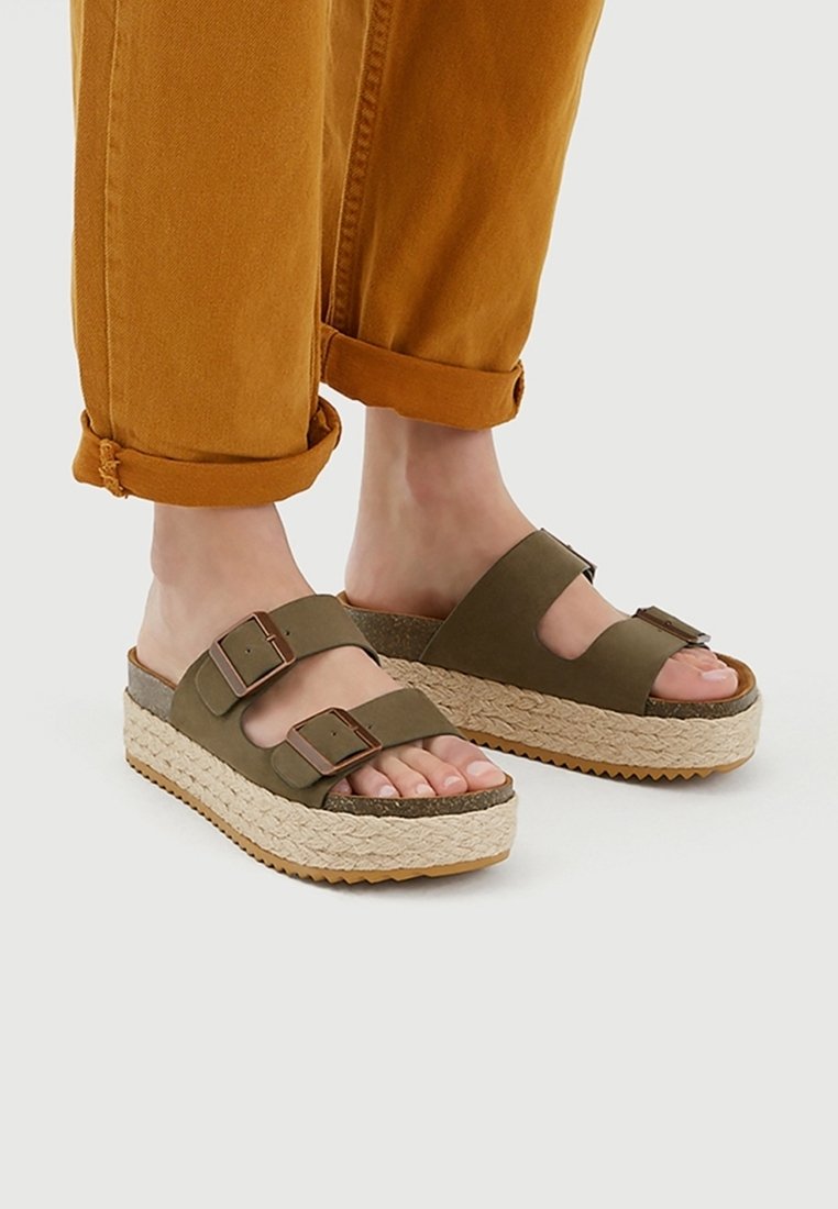 Pieds portant des sandales à plateforme vert olive avec des brides à double boucle, assorties à un pantalon moutarde retroussé sur un fond clair.