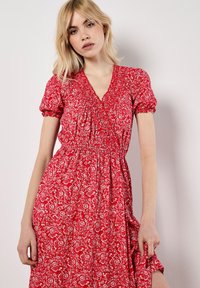 Robe florale rouge avec de courtes manches bouffantes, décolleté en V et taille froncée. Le tissu présente un motif floral blanc sur un fond vibrant.