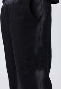 Primer plano de pantalones negros con bolsillos y diseños en forma de estrella cosidos en el muslo, contra un fondo liso.