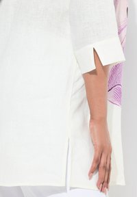 Tunique en lin blanche avec une fente latérale, ornée d'un motif abstrait violet et de manches mi-longues avec un petit détail découpé.