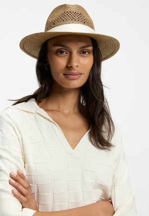 Vrouw met lang donker haar die een geweven tan fedora met witte band en een witte getextureerde V-hals top draagt, armen gekruist tegen een effen achtergrond.