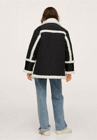 Sort polstret frakke med hvid fake pels, der har en bred krave og manchetter. Båret med lyseblå jeans og beige sko.