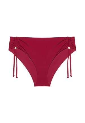 Mujer con cabello castaño ondulado que lleva un bikini rojo de dos piezas, de pie con una mano en la cadera frente a un fondo liso.