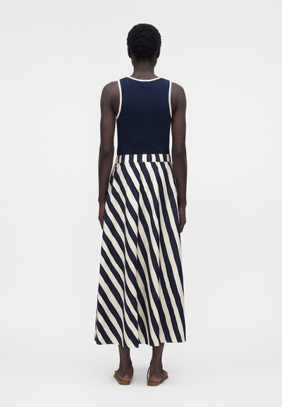 Άτομο που φοράει ένα σκούρο μπλε αμάνικο τοπ και μια ψηλόμεση, μακριά μέχρι τους αστραγάλους φούστα με διαγώνιες σκούρες μπλε και λευκές ρίγες, στέκεται μπροστά σε απλό φόντο.