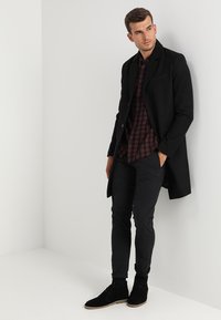 Cappotto di lana nera sopra una camicia a quadri bordeaux e nera, abbinato a pantaloni neri e stivaletti in suede. Il modello posa contro un muro bianco.