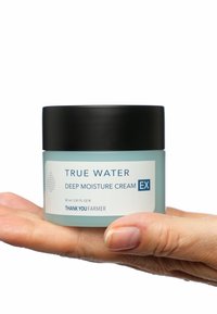 En liten, cylindrisk burk med ljusblå kräm med ett svart lock märkt "True Water Deep Moisture Cream EX" hålls i en hand.