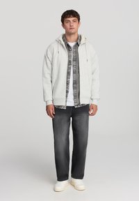 Felpa grigia chiara con un interno peloso, abbinata a una maglietta a righe bianco e nero, jeans scuri e sneakers bianche.